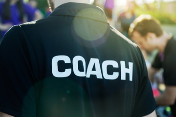sport coach in black shirt with white coach text on the back standing outdoor at a school field, wit
