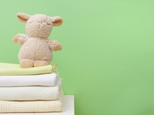  Stack Of Folded Up Natural Beige Cotton Blanket For Newborn And Decorative Toy Lamb On Green Background.