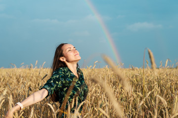 Wall Mural - A free woman on a wheat field with her hands open breathes air against the background of a rainbow, freedom, ecology