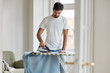 © WHstudio Leushin N - Hard working young Caucasian bearded husband irones clothes on ironing board, does domestic duties while wife is away, stands in living room, being busy with work about house. Chores concept