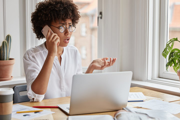 Indignant freelancer has serious telephone conversation, troubles with work, tries to solve problem, surrounded with papers, drinks hot beverage, looks desperately into distance, holds smart phone