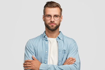 Horizontal shot of handsome self confident businessman or designer, stands crossed hands against white background, wears casual shirt, poses against white background. People and lifestyle concept