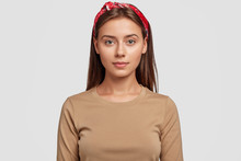 Portrait Of Pleasant Looking European Brunette Girl Wears Stylish Red Headband, Beige Sweater, Looks Confidently Directly In Camera, Listens Attentively Interlocutor, Poses Indoor. Style Concept