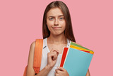 Fototapeta  - People, studying and university concept. Puzzled clever intern listens attentively lecture, holds colourful folders, carries rucksack, stands against pink background, purses lips in bewilderment.