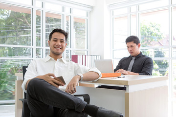 Happy young businessman sitting in the office. Business on mobile phone and computer. Laptop on the table.