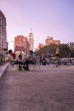 Diverse People Seated At Public Tables In Madison Square Park Area Of New York City With Empire State Building In Background