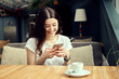 © photoguns - Young woman sitting in coffee shop at wooden table, drinking coffee and using smartphone. White cup of espresso stands on table. Girl holding phone, browsing internet, chatting, blogging, texting.