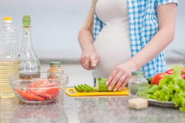 Close up pregnant woman with knife on kitchen cuts cucumber