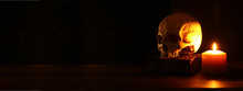 Human Skull, Old Book And Burning Candle Over Old Wooden Table And Dark Background.