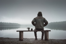 A man is sitting on a bench and looking at the lake. Back view. Also a coffee maker and a mug on the bench.