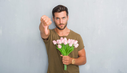 Sticker - Handsome young man over grey grunge wall holding flowers bouquet with angry face, negative sign showing dislike with thumbs down, rejection concept