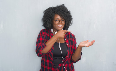 Canvas Print - Young african american woman over grey grunge wall wearing headphones amazed and smiling to the camera while presenting with hand and pointing with finger.