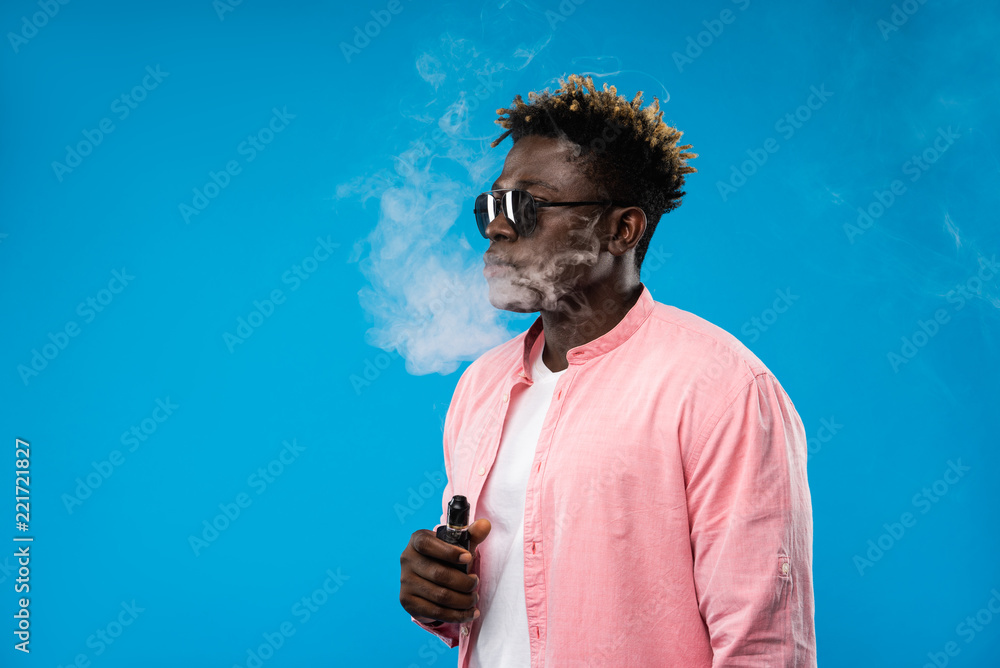 holding in vape smoke My addiction. Side view portrait of guy device and exhaling cloud of smoke. He is wearing pink shirt and sunglasses. Isolated on blue background. Copy space left side Stock Best Dry Herb Vaporizers tax-eliquid Colour:Aurora Pink with Pink Lemonade