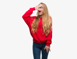 Sticker - Blonde teenager woman wearing red sweater peeking in shock covering face and eyes with hand, looking through fingers with embarrassed expression.