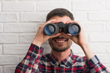 Young adult man over brick wall looking through binoculars with a happy face standing and smiling with a confident smile showing teeth