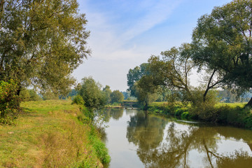 Wall Mural - Small river on the background of grass-covered banks against blue sky. River landscape on a autumn morning