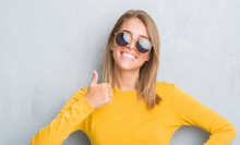 Beautiful Young Woman Standing Over Grunge Grey Wall Wearing Retro Sunglasses Happy With Big Smile Doing Ok Sign, Thumb Up With Fingers, Excellent Sign