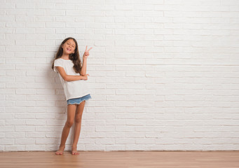 Wall Mural - Young hispanic kid stading over white brick wall smiling with happy face winking at the camera doing victory sign. Number two.