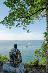 Wall Mural - Rear view of young man sitting at coast overlooking the ocean wearing colorful african shirt, Bubaque, Guinea Bissau