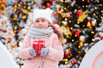 Wall Mural - happy child girl holding christmas gift outdoor on the walk in snowy winter city decorated for new year holidays. Trees with christmas lights on background