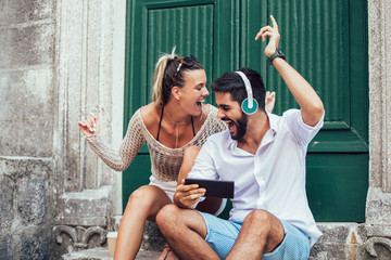 Portrait of beautiful smiling young tourist couple sitting on stairs using tablet and listening to music.