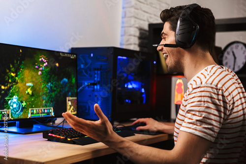 Fotomural Image of cheerful gamer man playing video games on computer, wearing headphones