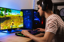 Image Of Young Man Playing Video Games On Computer, Wearing Headphones And Using Backlit Colorful Keyboard