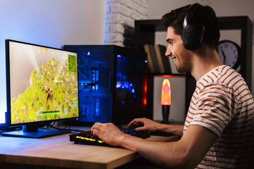 Wall Mural - Portrait of european gamer guy playing video games on computer, wearing headphones and using backlit colorful keyboard