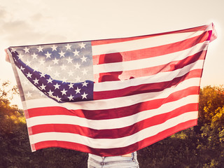 Attractive man holding a US flag against the background of the rays of the setting sun. View from the back. Preparing for the holidays