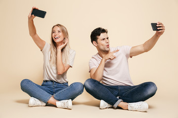 Poster - Young loving couple sitting isolated over beige wall background make selfie by mobile phones.