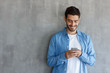 © Damir Khabirov - Indoor picture of European Caucasian man isolated on gray background dressed in blue denim shirt and typing messages on his smartphone communicating with friends in social networks, smiling happily