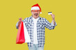 © khosrork - Suprised open mouth middle aged man in casual style and new yar red hat, standing, holding shopping bags and credit card with amazed face, looking at camera. Studio shot, isolated on yellow background
