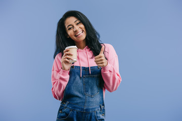 Sticker - smiling young african american woman doing thumb up and holding disposable coffee cup isolated on blue background