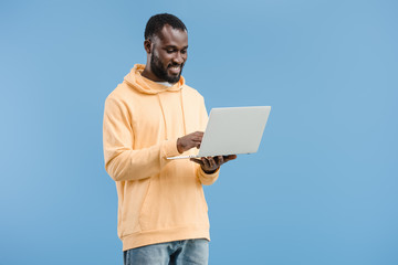 smiling young african american man using laptop isolated on blue background