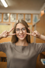 Smiling young student schoolgirl lady with long hair wearing eyeglasses standing in empty classroom looking camera showing peace gesture.