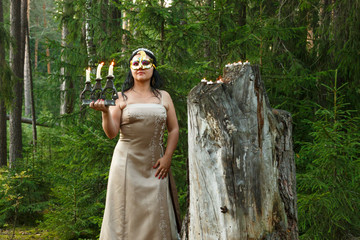 Forest fairy in a light dress with a mask on her face in the forest with a candlestick with candles.