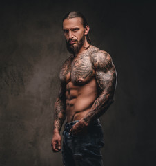 Wall Mural - Portrait of a shirtless bearded tattooed male posing with hand in pocket. Isolated on dark textured background.