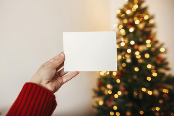 christmas mockup with space for text. hand holding empty greeting card on background of golden beautiful christmas tree with lights in festive room. cozy winter holidays..