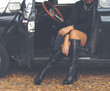 Sexy lady sitting in a classic car. Long leather boots and black dress. Image has a vintage effect applied.