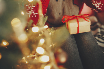 Christmas gift box on girl legs at golden beautiful christmas tree with lights in festive room. winter holiday atmospheric moments. seasons greetings. Merry Christmas