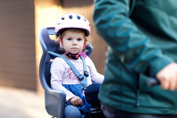 Portrait of little toddler girl with security helmet on the head sitting in bike seat and her father or mother with bicycle. Safe and child protection concept. Family and weekend activity trip.