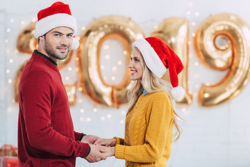 Wall Mural - smiling couple in santa hats holding hands at home with 2019 new year golden balloons