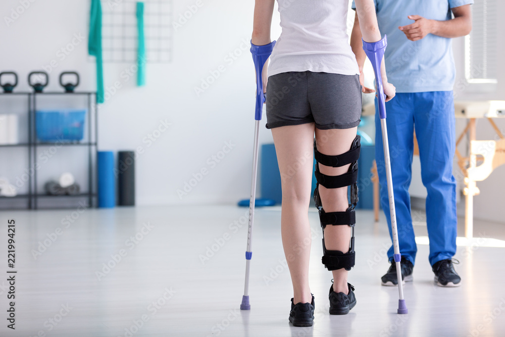 Patient with stiffener on the leg walking with crutches during rehabilitation - obrazy, fototapety, plakaty 