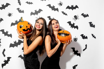 Two charming brunette women in black dresses with red lipstick hold Halloween pumpkins on a white background with bats and spiders