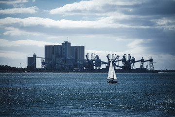 Small idyllic single sailing boat with white sail in blue water of sea on background of industrial manufacture on shore