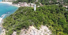 Aerial Shot Pulling Up And Away From A White Lighthouse In The Middle Of A Forested Hill Above A Quaint Beach Town.