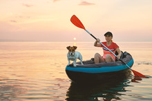 Red-haired Young Woman Is Rowing On An Inflatable Kayak By The Sea With A Dog Jack Russell Terrier On A Background Of Pink Sunrise In Beautiful Nature. Great Disk Of The Rising Sun. Sun Rays. Sport