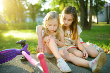 Adorable girl comforting her little sister after she fell off her scooter at summer park. Child getting hurt while riding a kick scooter.