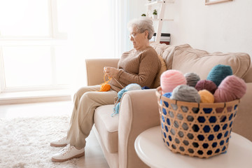 Wall Mural - Senior woman sitting on sofa while knitting sweater at home