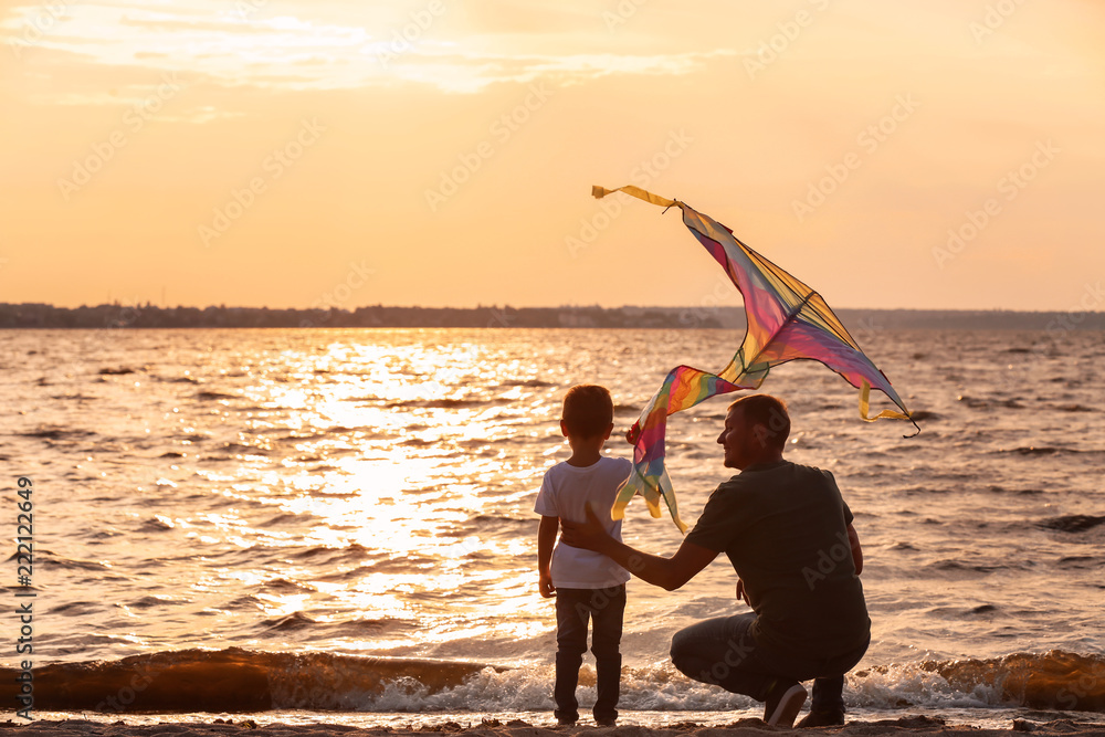Happy father and son flying kite near river at sunset